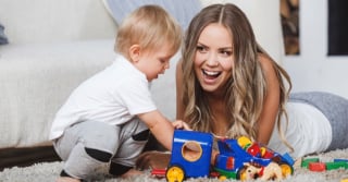 Young woman plays with small child on carpeted floor.  Many toys are scattered about