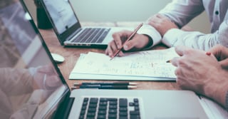 Reviewing a document on a table that has 2 laptops set up to either side of the document