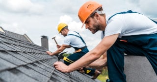 Men in hard hats using hammers to secure shingles to a roof