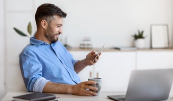 white male small business owner looks at phone while sitting at desk with computer and coffee