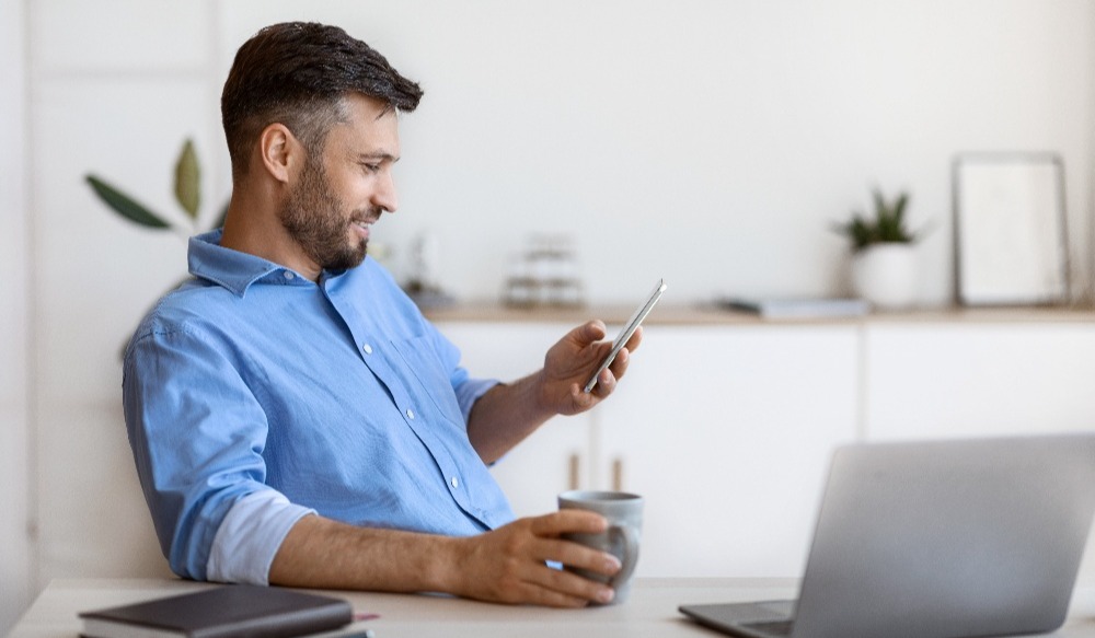 white male small business owner looks at phone while sitting at desk with computer and coffee